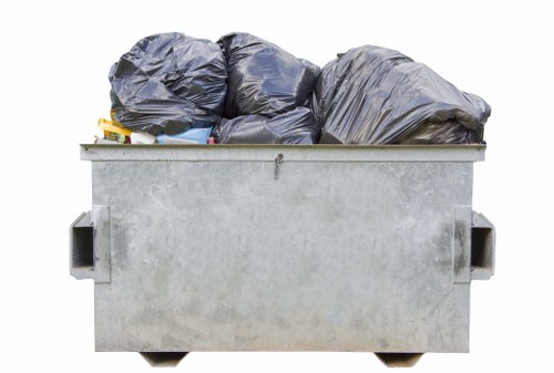 The image shows a large grey metal commercial waste skip filled with black plastic rubbish bags stacked on top of each other. The bags appear to be of varying sizes and are tightly sealed, with some exhibiting a slightly crinkled texture. The skip is positioned outdoors on a paved surface, likely on a commercial or industrial premises, with the background suggesting an open environment typical of waste disposal areas. The skip’s sturdy steel construction and rectangular shape are clearly visible, with handles on each side for maneuvering. The environment appears well-lit, possibly by natural daylight, highlighting the metallic finish of the skip and the dark color of the rubbish bags. This setup exemplifies typical waste collection practices in areas like Spitalfields, where commercial waste removal services, such as those provided by Commercial Waste Removal Spitalfields, are employed to manage large quantities of refuse efficiently, aligning with local waste management needs in the central London borough.