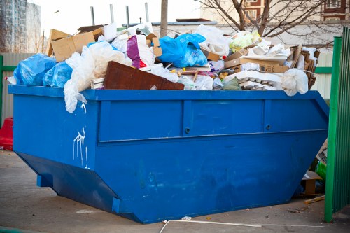 A large blue industrial skip filled with various types of waste, including white plastic bags, cardboard boxes, and loose paper, positioned on a paved surface in an urban outdoor setting. The skip, made of metal with a smooth finish, is overflowing with rubbish, some items spilling over the top edges. It is situated near a green metal fence, and in the background, there are leafless trees and buildings, suggesting a cold season. The scene appears to be part of a waste collection or rubbish removal operation, with the surrounding environment indicating a residential or commercial area in Spitalfields. The lighting is natural, with diffuse daylight illuminating the scene, emphasizing the textures of the waste materials and the bright blue colour of the container, supporting the services offered by Commercial Waste Removal Spitalfields for rubbish disposal and waste management.