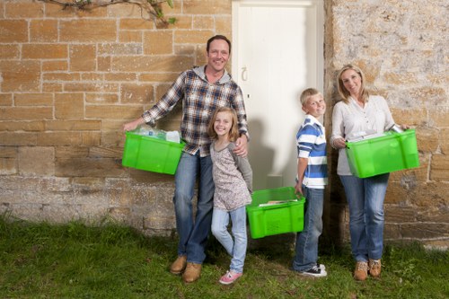 A family of four, including a man, woman, and two children, are standing outdoors in front of a weathered stone and brick wall, each holding bright green plastic recycling bins filled with waste items. The man, positioned on the left, is smiling and wearing a casual checked shirt and jeans, holding a bin in his right hand. The young girl in front of him is wearing a patterned long-sleeve top and jeans, smiling at the camera, with a bin in her left hand. To the right of the girl, a boy in a striped shirt and jeans stands beside a woman, who is also smiling and wearing a white blouse with jeans, holding a green bin. The scene suggests an activity related to rubbish collection or recycling, set on a patch of grass with a subtle natural atmosphere. The background features an aged stone wall with visible signs of weathering, typical of architectural features around Spitalfields, London, associated with rubbish and waste management services offered by Commercial Waste Removal Spitalfields, focusing on efficient waste disposal and recycling.