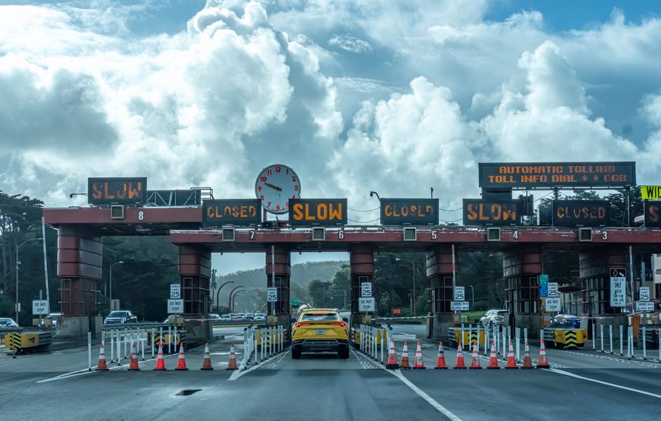 A busy toll booth with multiple lanes, each separated by red cylindrical pillars with metallic finishes. Several orange and white traffic cones direct vehicles through the lanes, some of which display