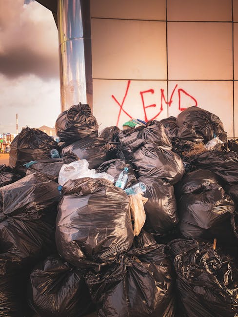 A green recycling bin designed for collecting old clothing items is positioned outdoors against a background of foliage and waste debris. The bin has a rectangular shape with a slightly sloped roof an