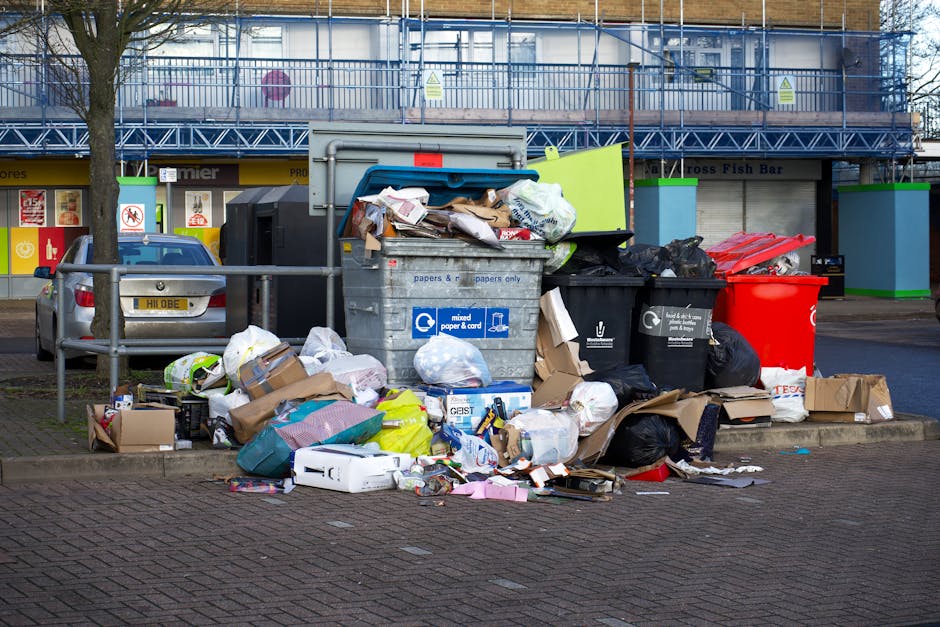 A row of large outdoor waste bins positioned along a pavement beside a brick wall, used for rubbish collection. The bins are made of durable plastic with textured lids, with the closest bin painted in