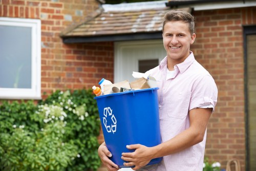 A young man with short brown hair and a smiling expression, wearing a light pink short-sleeved shirt, is standing outdoors in front of a brick house with a window and tiled roof. He is holding a blue recycling bin with a prominent white recycling symbol on the side, filled with various pieces of rubbish including cardboard, paper, plastic bottles, and other household waste. The environment features a paved driveway or garden path, with greenery and flowering plants visible in the background near the house, suggesting a suburban setting. The scene conveys waste disposal or rubbish removal activity typical to residential areas in Spitalfields, with the man ready to load or transport waste for collection by services such as Commercial Waste Removal Spitalfields.